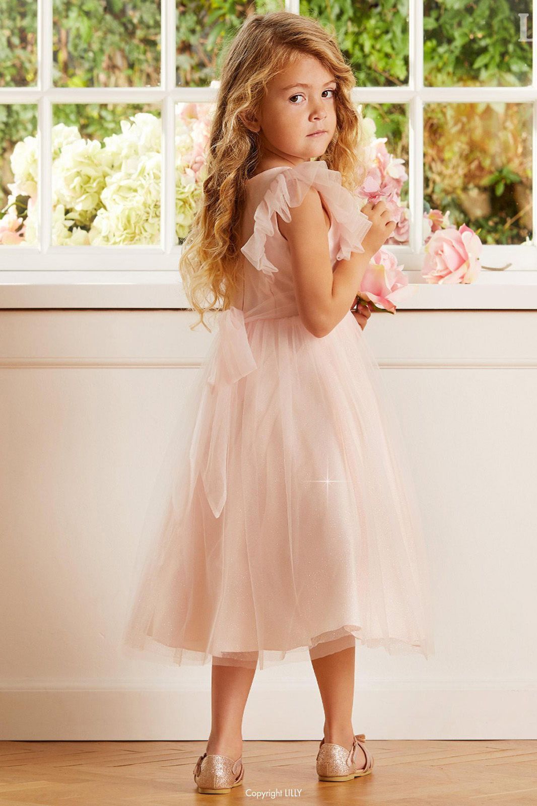 Ein kleines Mädchen in einem rosa Kleid steht mit einem Blumenstrauß in der Hand vor einem Fenster.