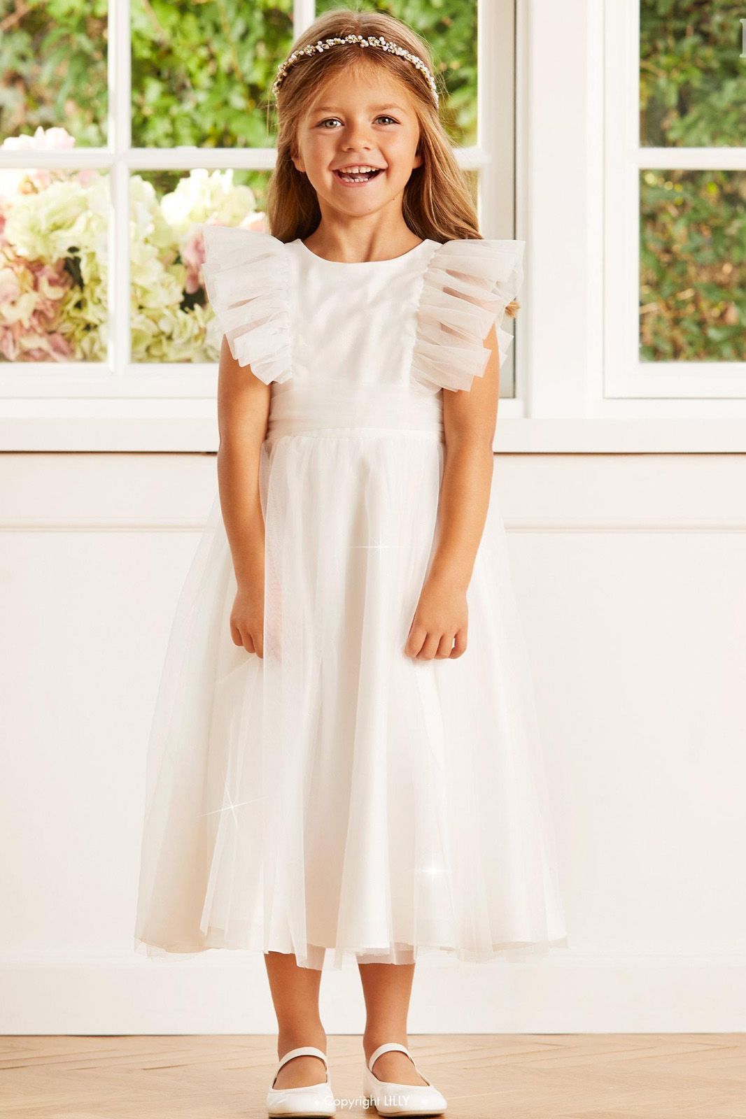 Ein kleines Mädchen in einem weißen Kleid steht vor einem Fenster.