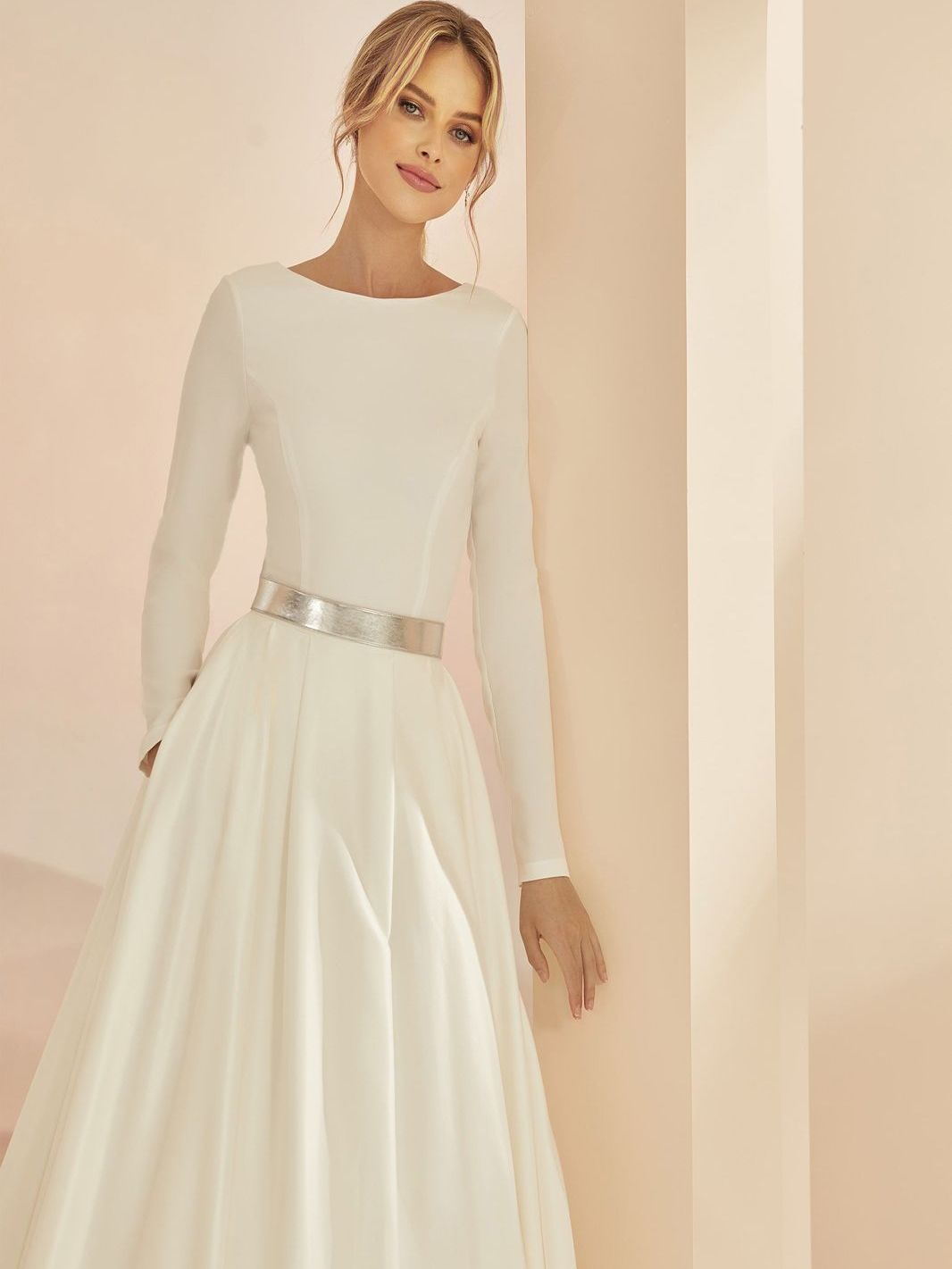 Eine Frau trägt ein weißes Brautkleid mit langen Ärmeln und einem silbernen Gürtel.