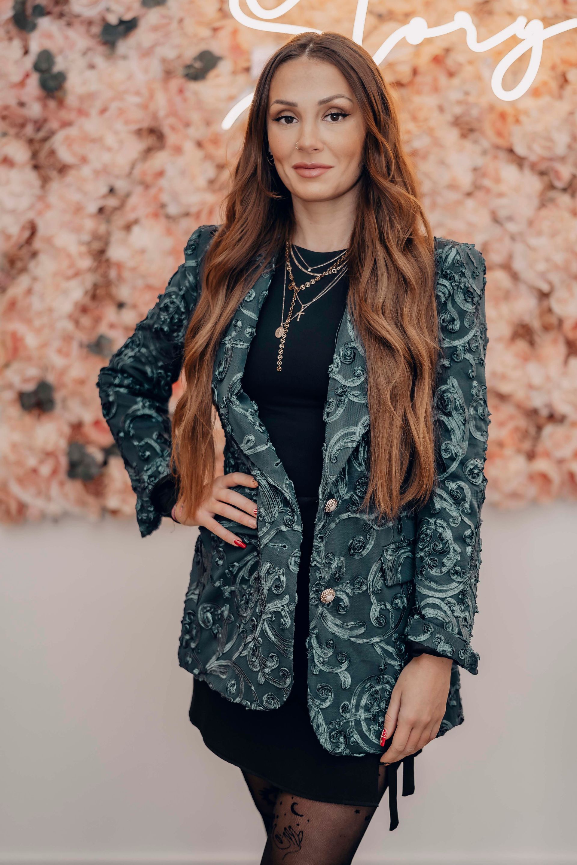 Eine Frau in einer grünen Jacke und einem schwarzen Kleid steht vor einer Blumenwand.