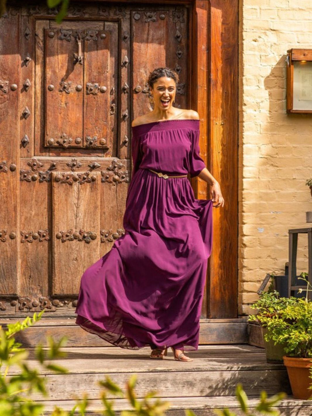 Eine Frau in einem lila Kleid steht vor einer Holztür.
