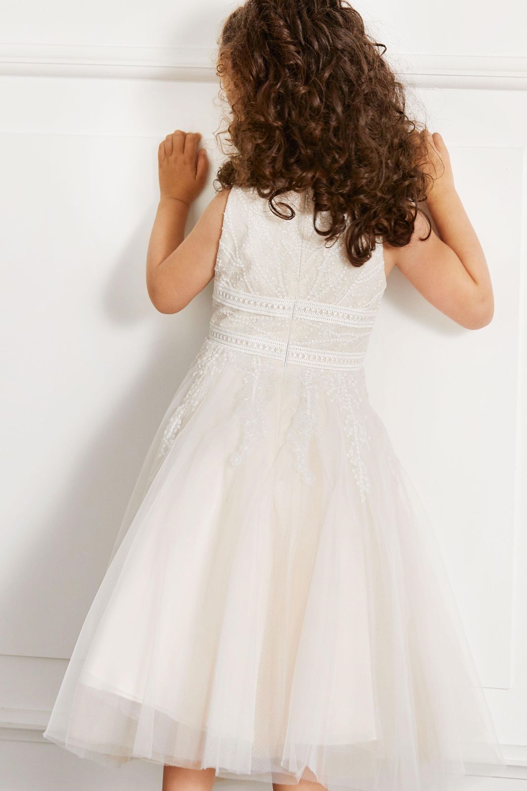 Ein kleines Mädchen in einem weißen Kleid lehnt an einer Wand.