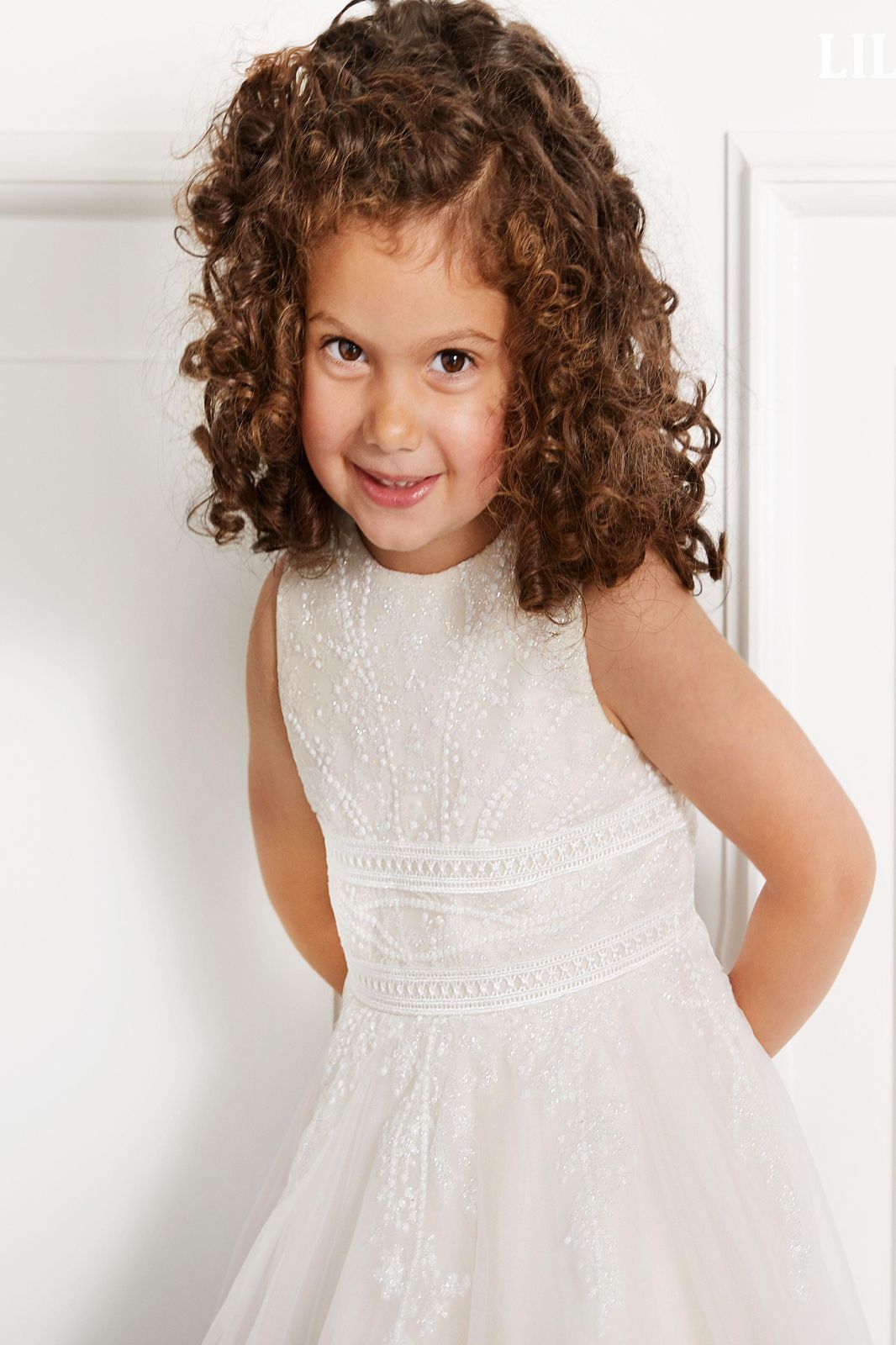 Ein kleines Mädchen mit lockigem Haar trägt ein weißes Kleid und lächelt.