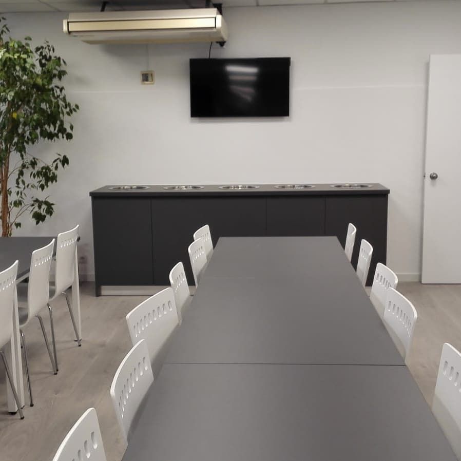 Sala de conferencias con mesas largas, sillas blancas, credenza negra, TV y árbol.