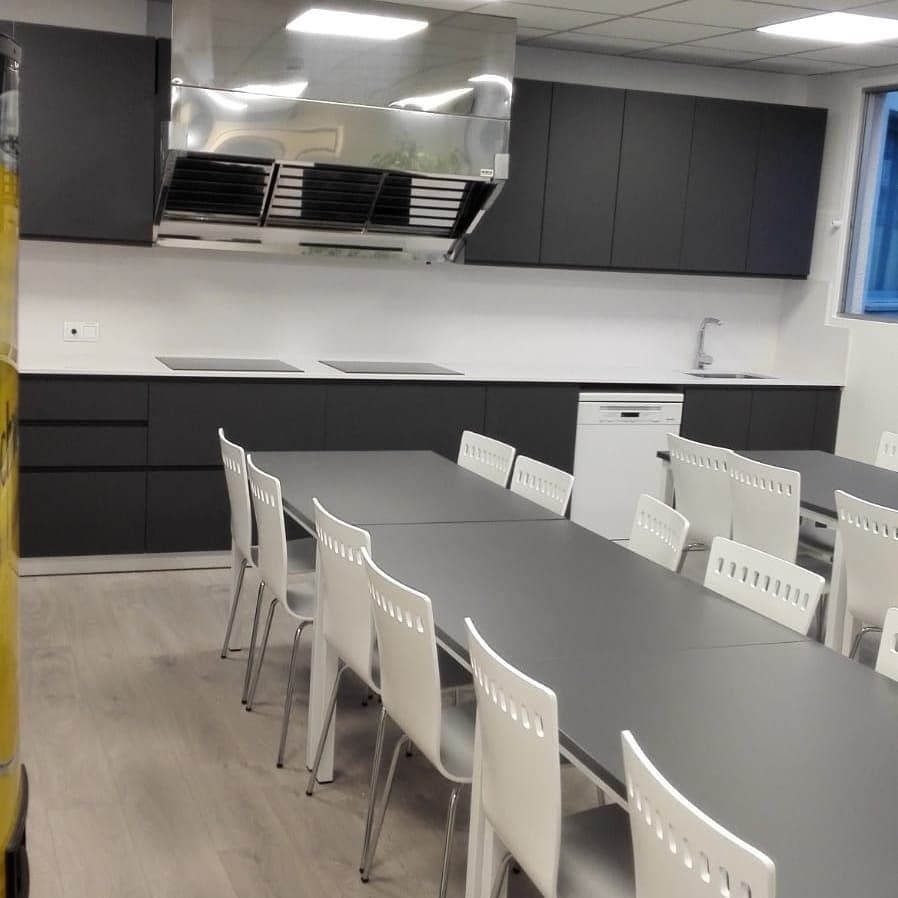 Cocina con mesas y sillas largas, gabinetes oscuros, campana extractora de acero inoxidable, paredes blancas.