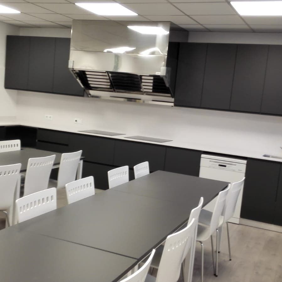 Aula de cocina con gabinetes negros, encimeras blancas, campana de acero inoxidable, mesas y sillas blancas.