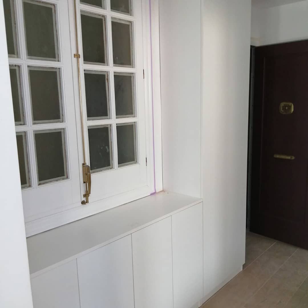 Ventana blanca con marco de madera, gabinetes blancos y una puerta marrón en un pasillo.