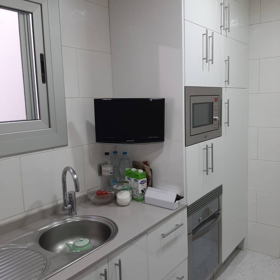 Cocina con gabinetes blancos, fregadero de acero inoxidable, microondas y pequeño televisor montado en la pared.