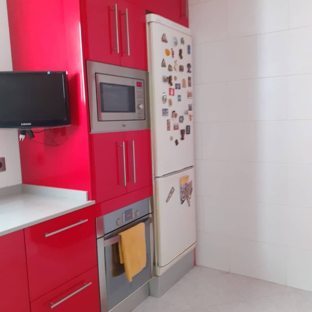 Muebles de cocina rojos con microondas, horno y refrigerador. Televisor de pared y encimera blanca.