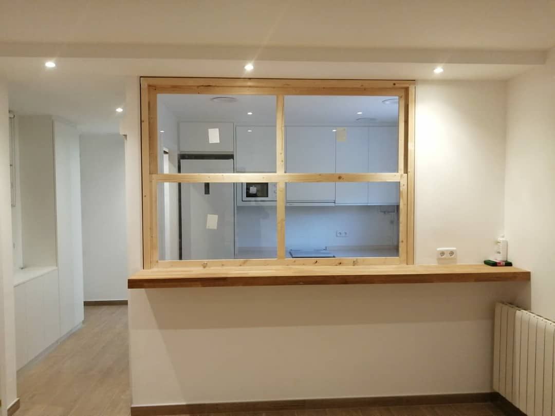 Vista interior de una ventana pasante abierta con marco y encimera de madera, frente a una cocina blanca.