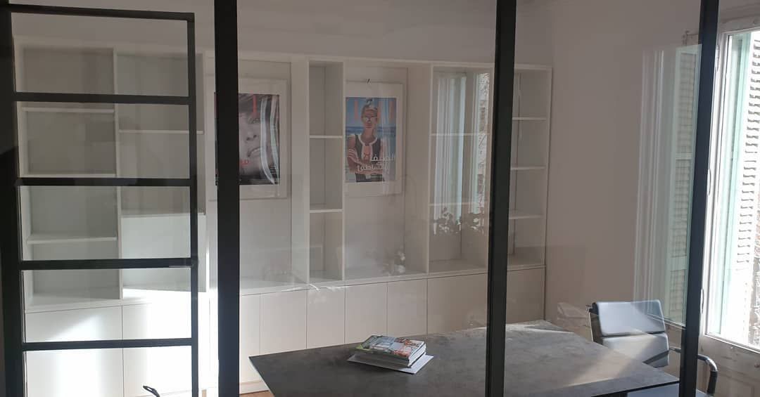 Interior de oficina con paredes de vidrio, estanterías y una mesa.