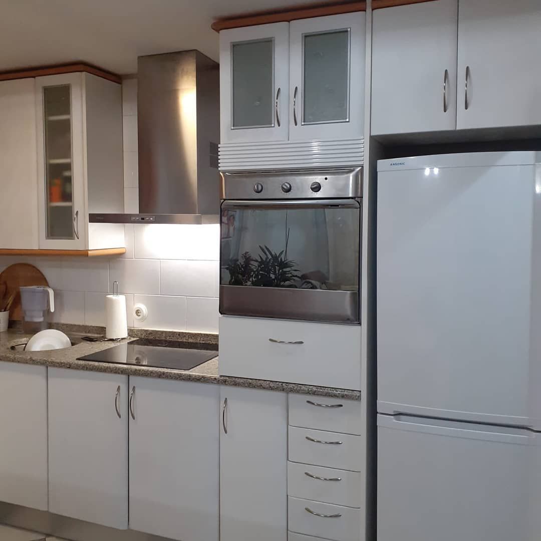 Cocina blanca con gabinetes, horno y refrigerador. Campana extractora de acero inoxidable y encimera moteada.