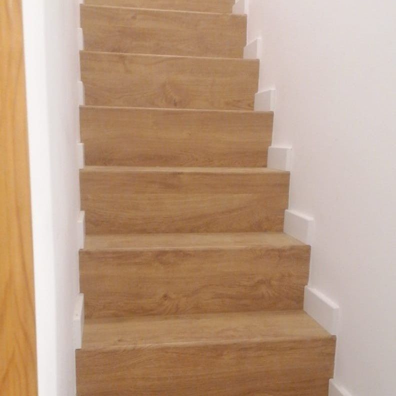 Escalera de madera con peldaños de color marrón claro y contrahuellas blancas contra una pared blanca.