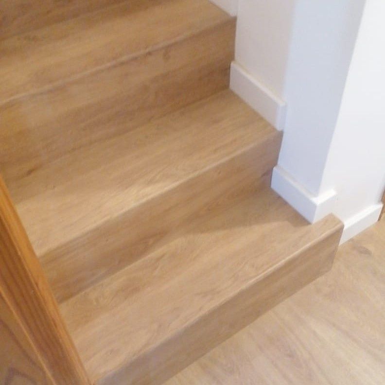 Escaleras de madera con peldaños y contrahuellas de color marrón claro, junto a una pared y molduras de color blanco.