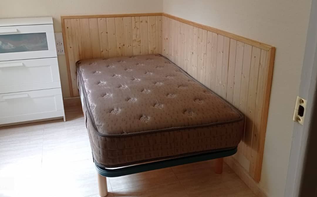 Colchón marrón sobre patas de madera en esquina, contra paredes revestidas de madera.