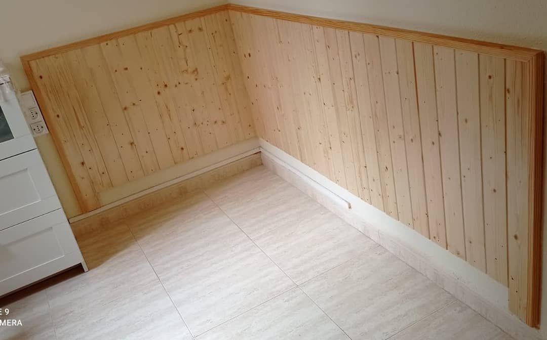 Revestimiento de pared de madera en un rincón de una habitación, con zócalos y suelo de color blanco.