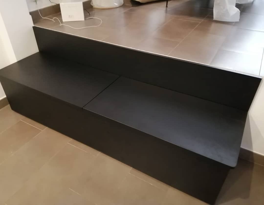 Banco negro con respaldo en una habitación, colocado sobre una plataforma elevada contra una pared.