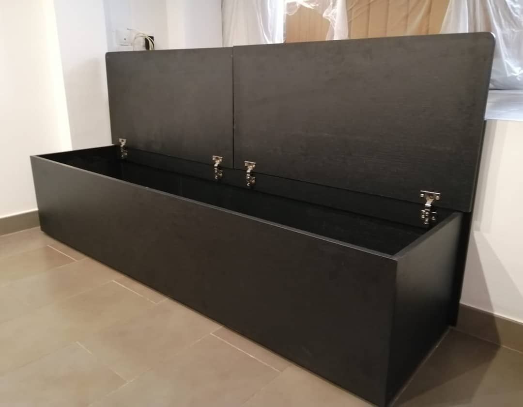 Banco de almacenamiento negro con tapa abierta, que revela un compartimento vacío, contra una pared blanca.