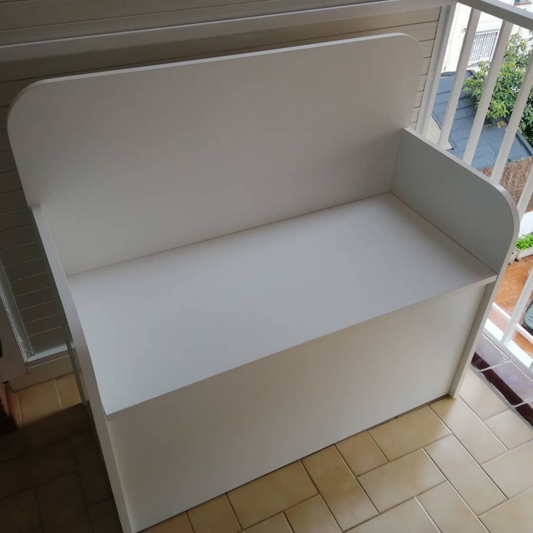 Banco de almacenaje blanco sobre balcón revestido de azulejos, con respaldo y paneles laterales curvados.