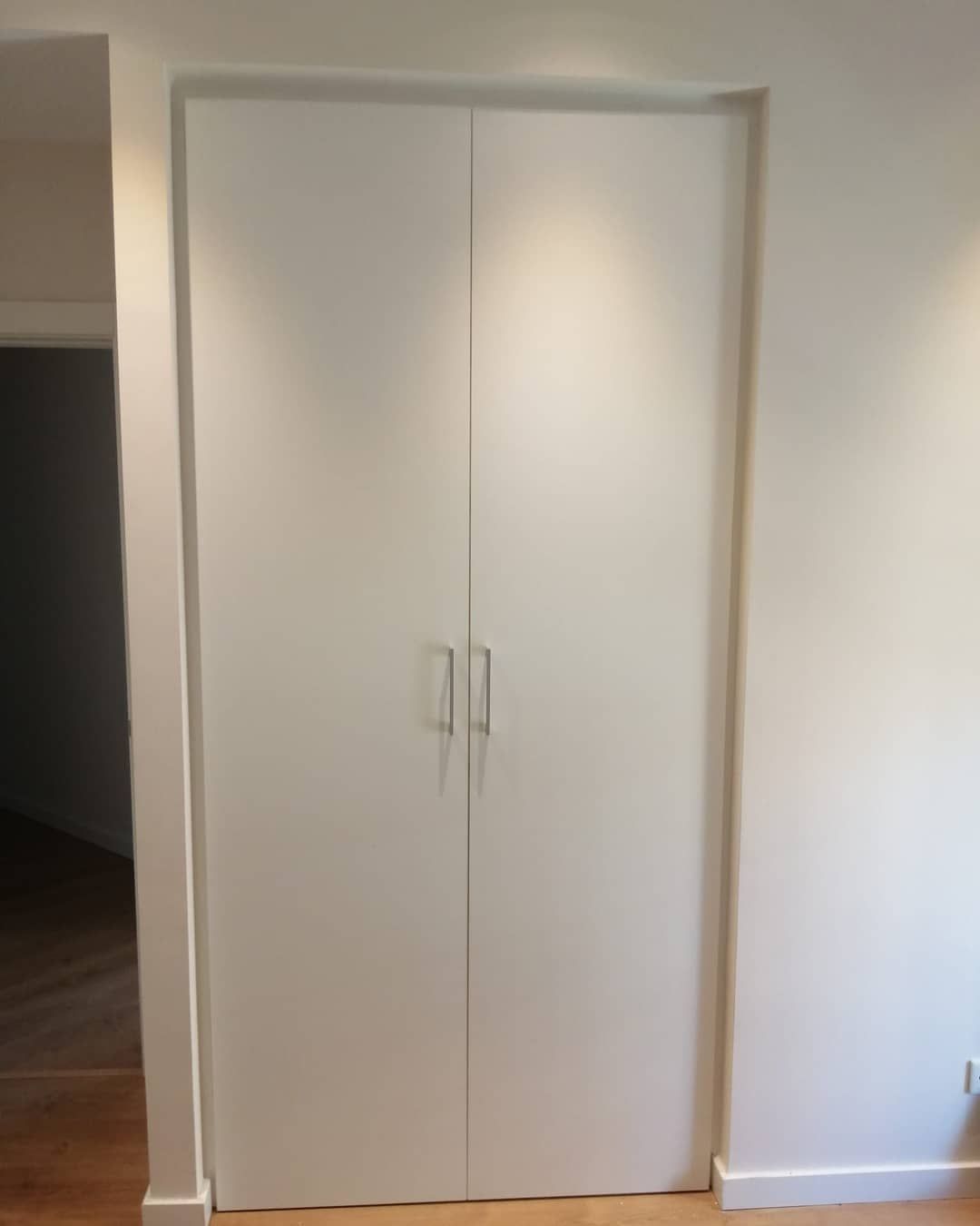 Puertas dobles blancas empotradas en una pared blanca con tiradores plateados minimalistas; iluminadas por luces de techo.