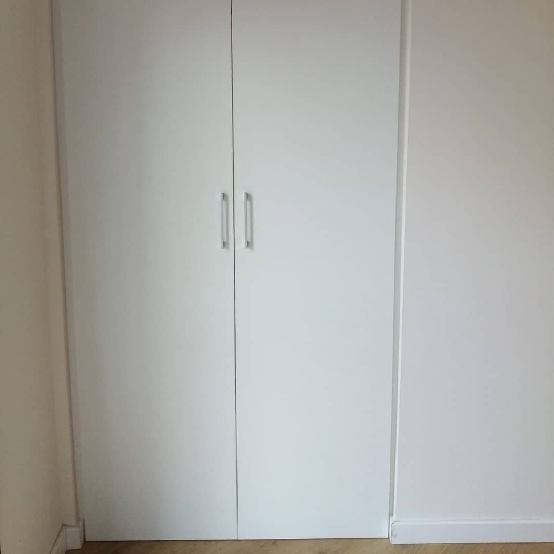 Armario blanco de doble puerta con dos tiradores plateados sobre paredes blancas y suelo de madera.