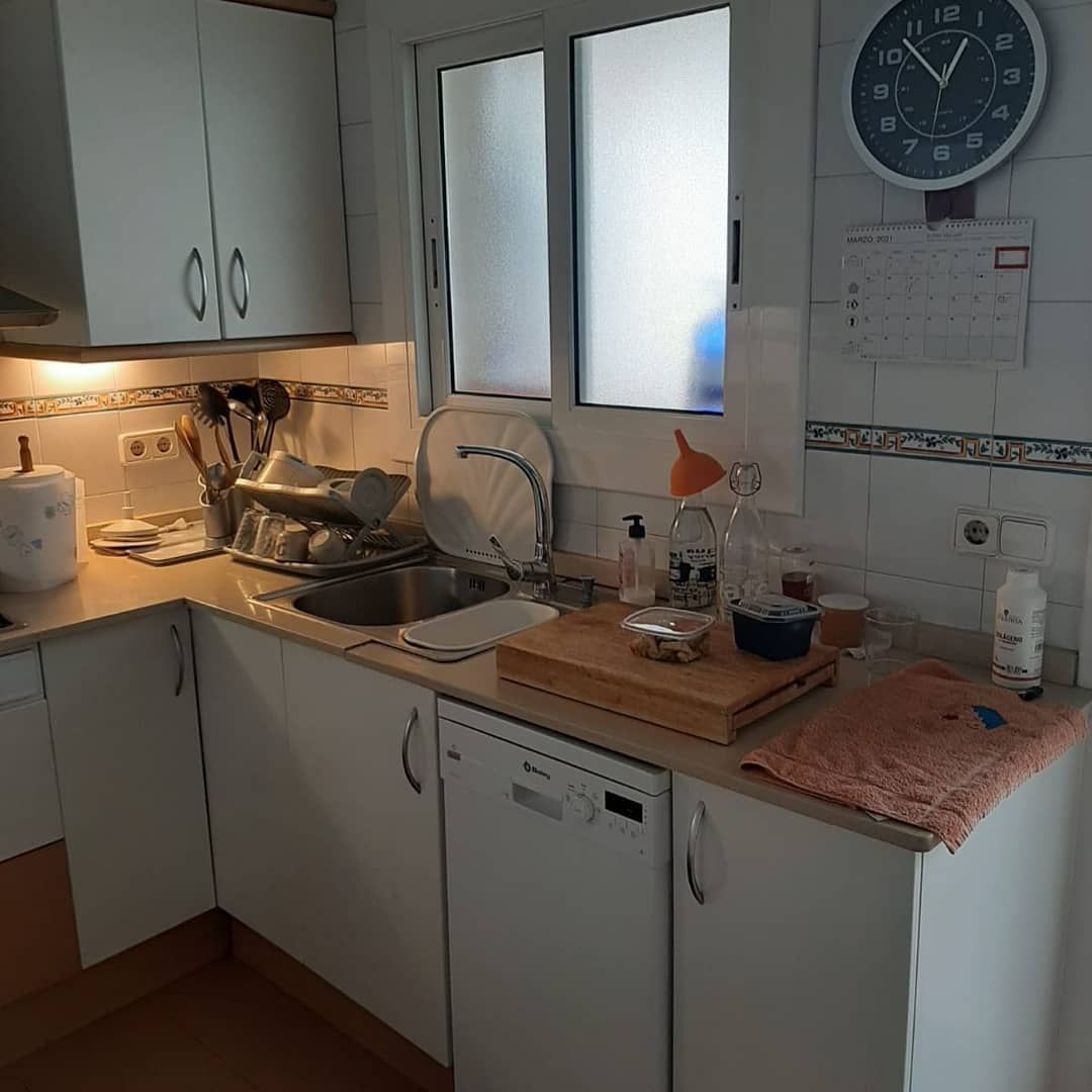 Una cocina pequeña con armarios blancos, una ventana y un reloj en la pared. Se ve un lavavajillas.