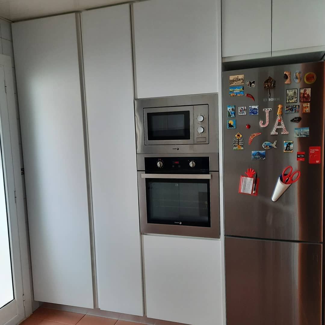 Muebles de cocina blancos con microondas y horno incorporados junto a un refrigerador de acero inoxidable.