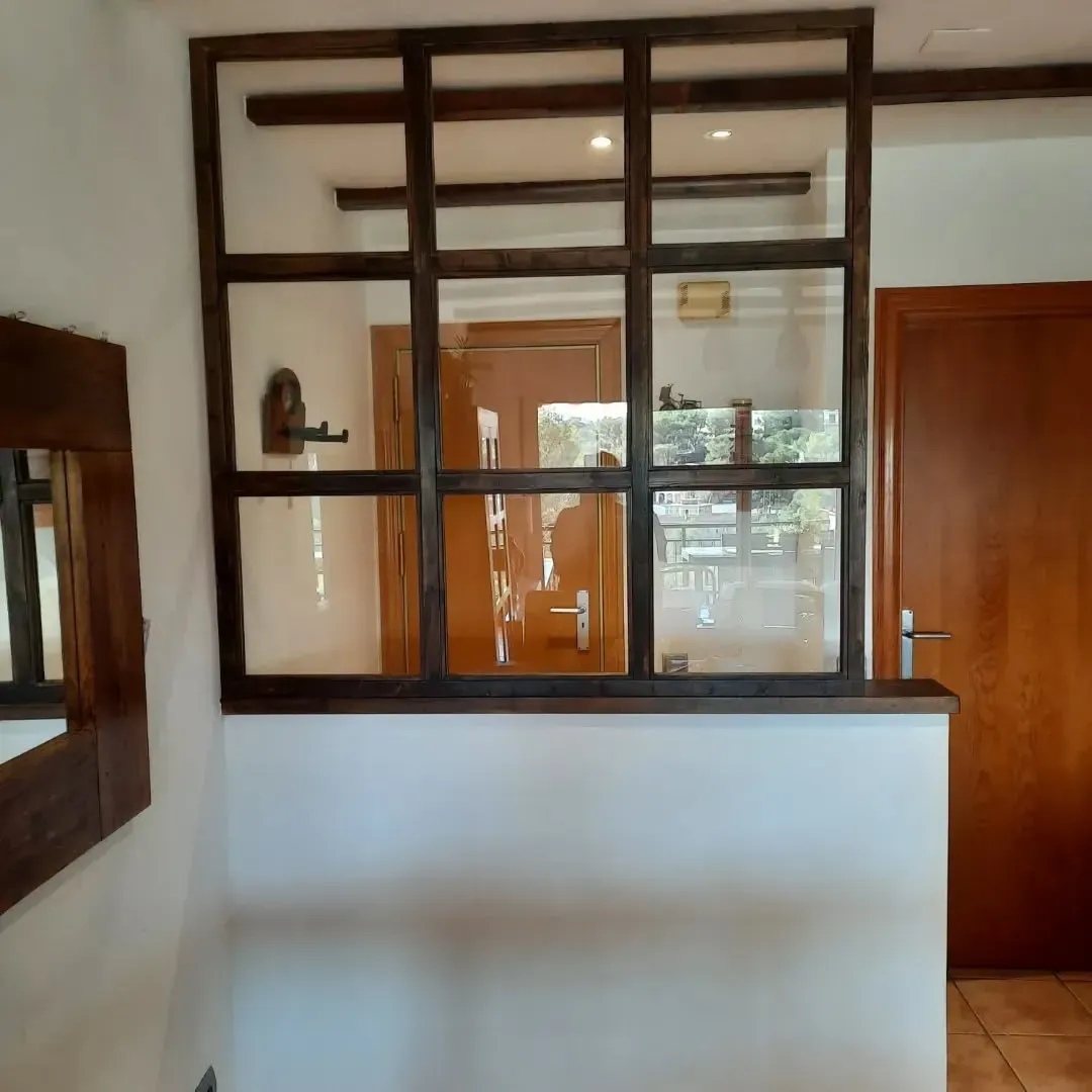 Ventana con marco de madera y mampara de vidrio, que separa dos espacios interiores con paredes blancas y una puerta.