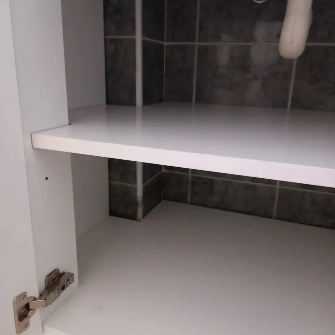 Mueble de baño blanco con puerta abierta, estante y fontanería visible.