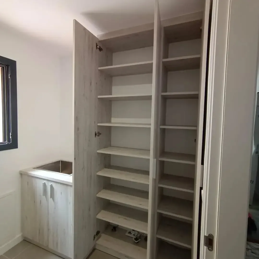 Armario alto de madera clara con puertas abiertas y estantes a la vista. A la izquierda hay una base para fregadero.