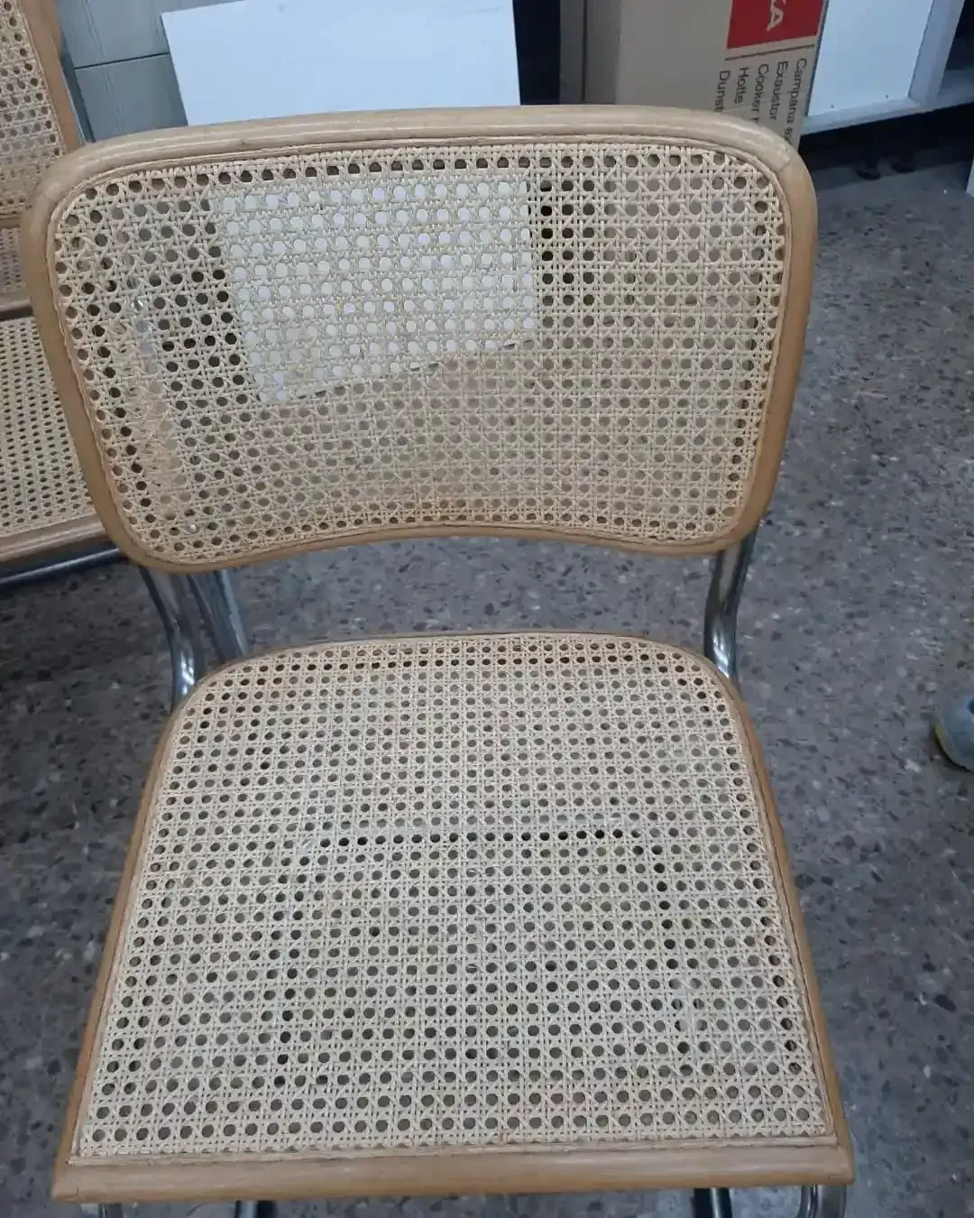 Silla de caña con respaldo curvado y patas cromadas, situada en un espacio interior indefinido.