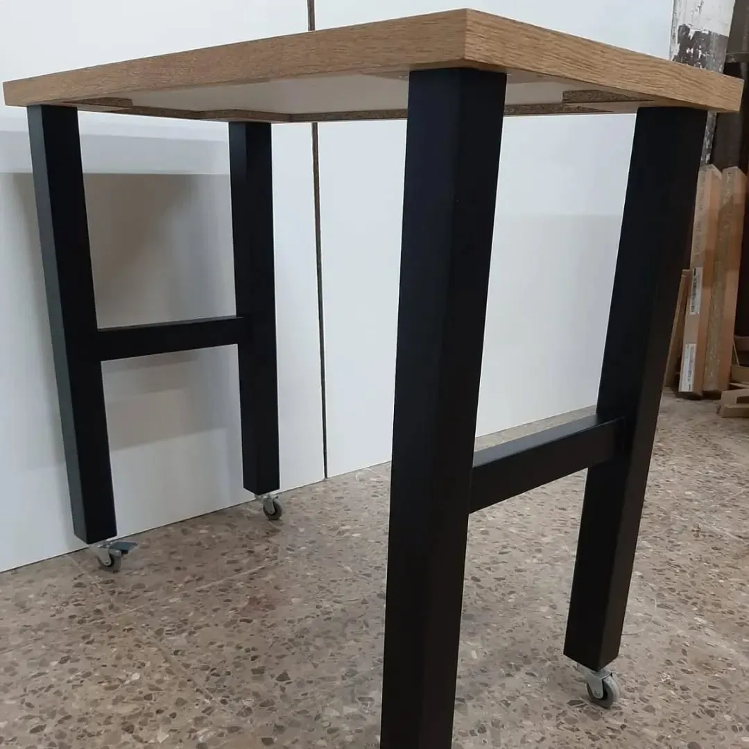 Mesa de trabajo de metal negro con ruedas, tapa de madera y patas en forma de H, para interior.