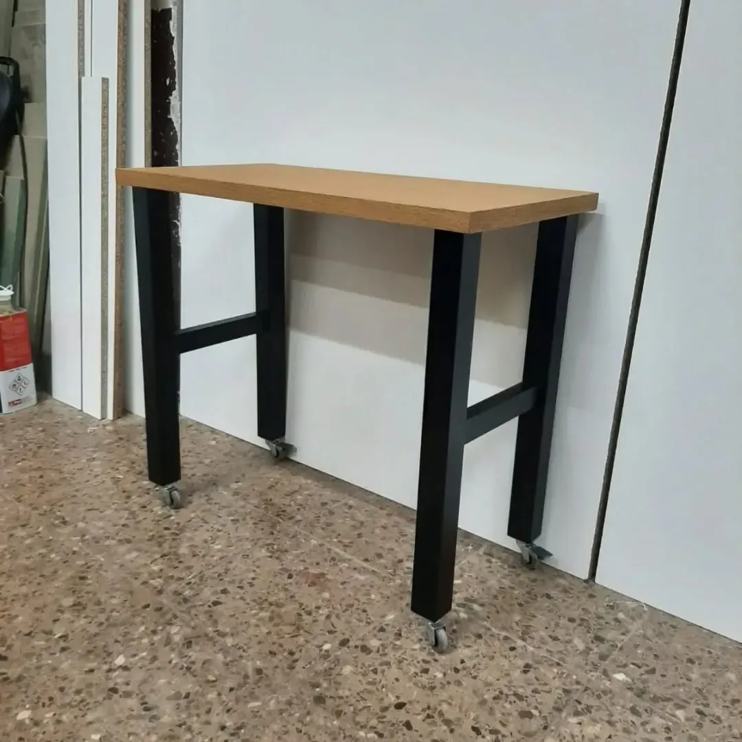 Pequeña mesa rectangular de madera con patas de metal negro, sobre ruedas, contra una pared blanca.