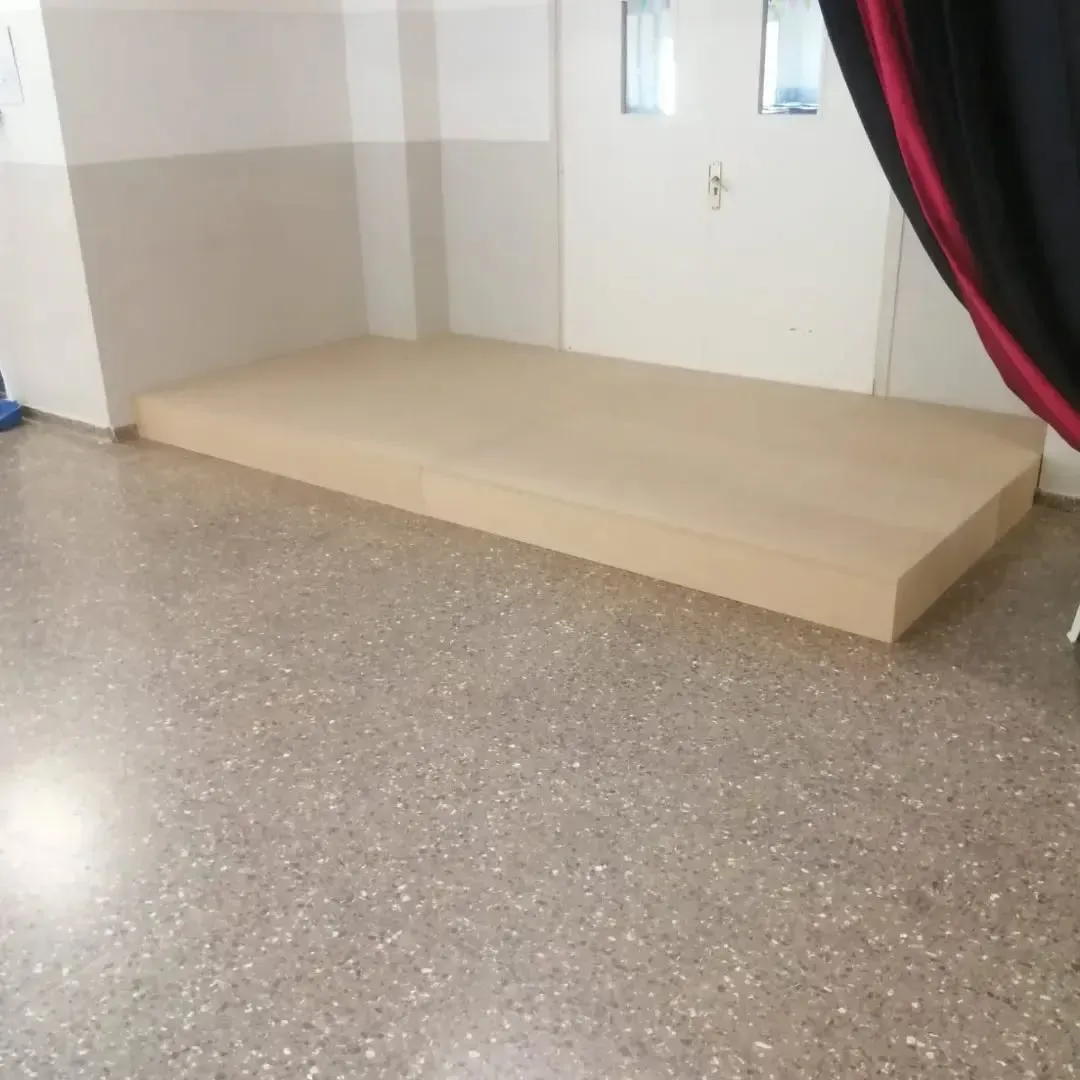 Una plataforma marrón claro a ras de suelo en una habitación con suelo de terrazo. Al fondo se ve una puerta.