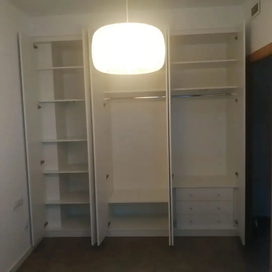 Armario blanco con puertas abiertas; estantes, barras de colgar y cajones visibles. Una lámpara cuelga encima.