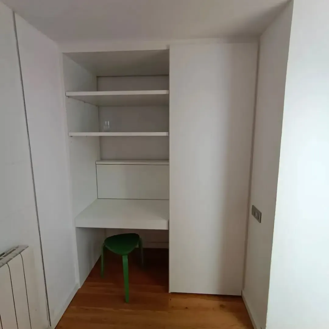 Escritorio blanco empotrado y estantería en una habitación con suelo de madera y un taburete verde.