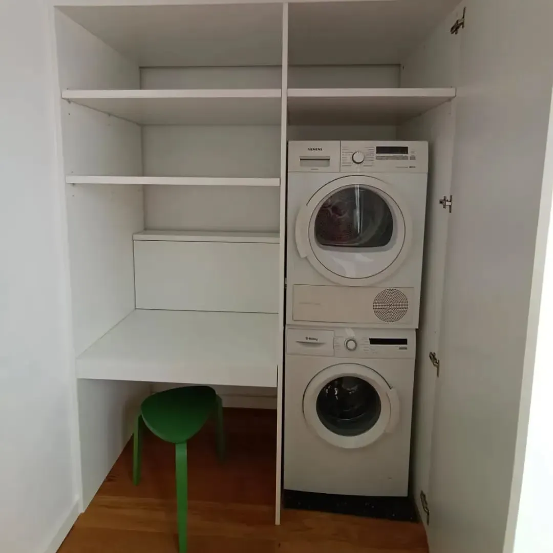 Mueble blanco con lavadora y secadora apiladas, estantes y un pequeño taburete verde.