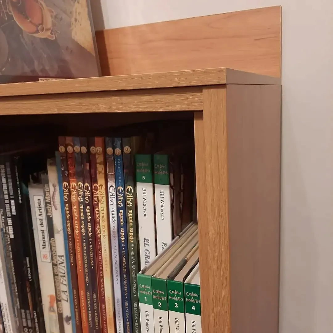 Estantería llena de libros, acabado en madera marrón, contra una pared blanca.