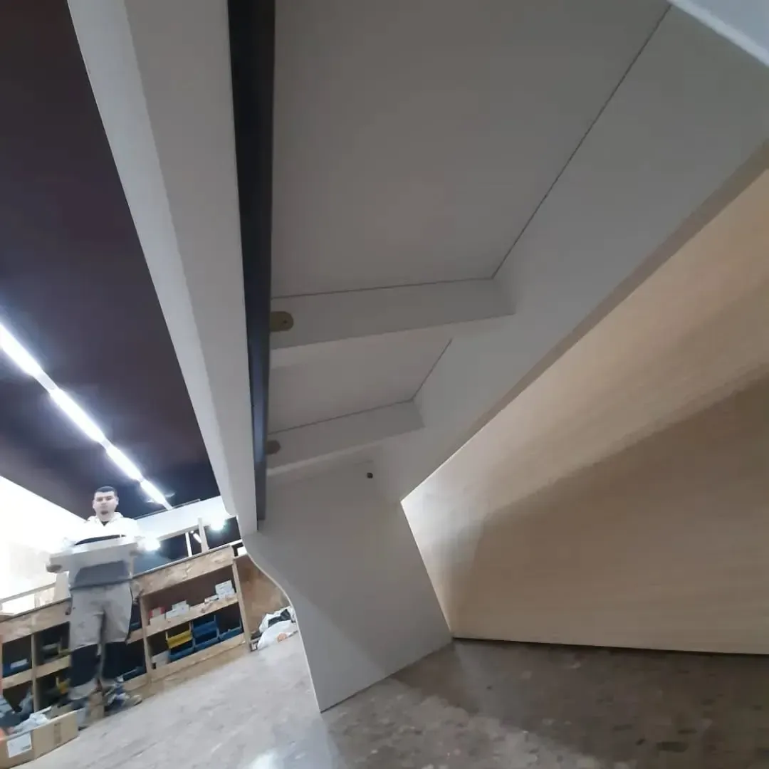 Una persona se encuentra bajo una estructura angular blanca en un taller. Panel de madera claro a la derecha.