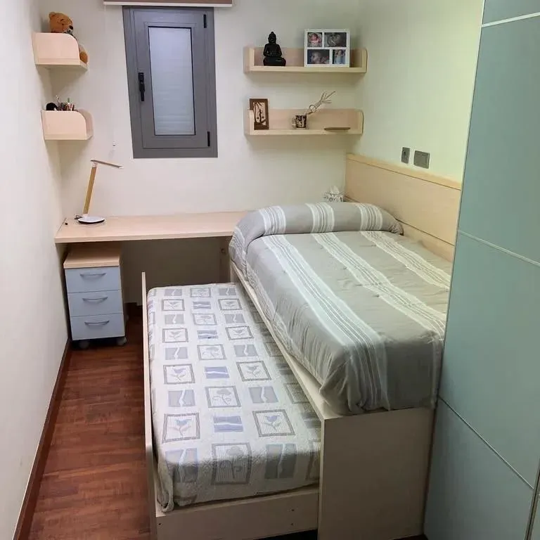 Dormitorio pequeño con cama plegable, escritorio, estanterías y ventana.