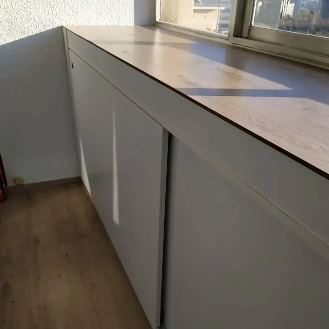 Armarios de almacenaje empotrados de color blanco con puertas corredizas bajo una encimera de madera, junto a una ventana.