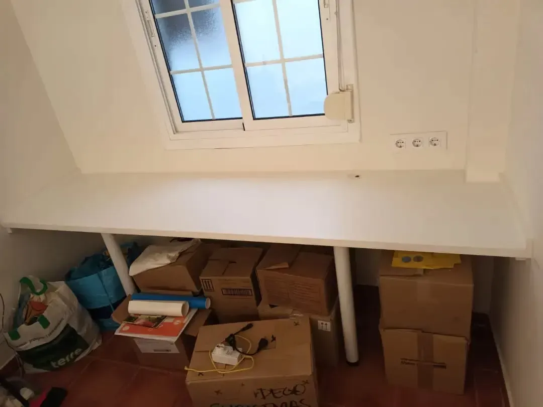 Estantería blanca empotrada bajo una ventana con varias cajas de cartón y elementos de almacenaje.