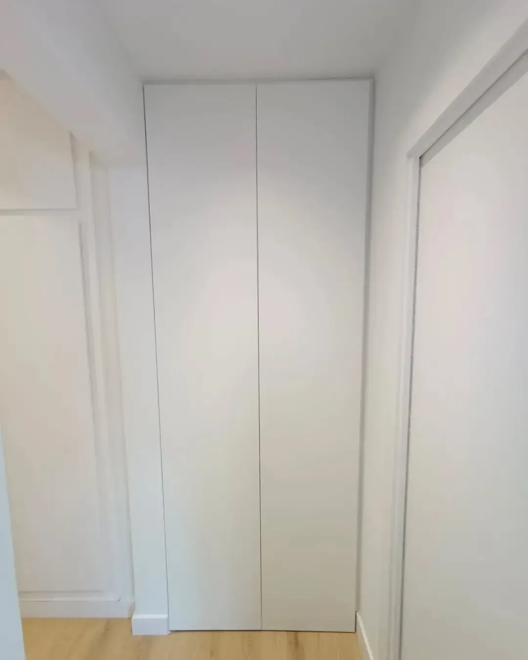Puertas de armario blancas en un pasillo estrecho. Suelo de madera clara y paredes blancas.