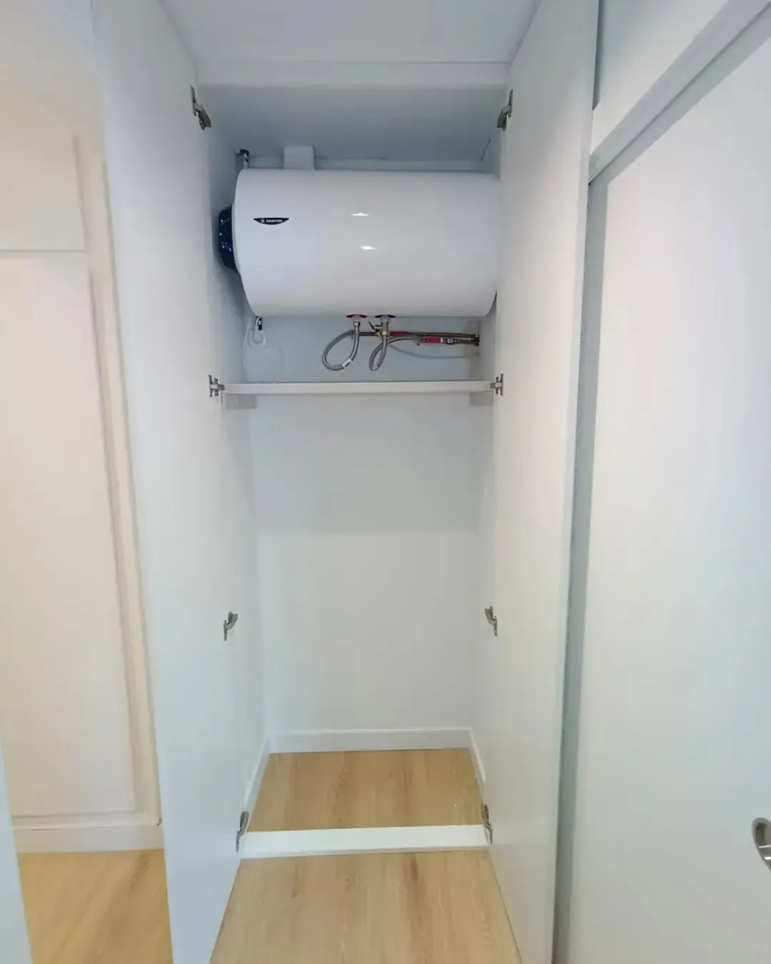 Armario blanco con calentador de agua montado en la parte superior; debajo hay una barra para colgar artículos.