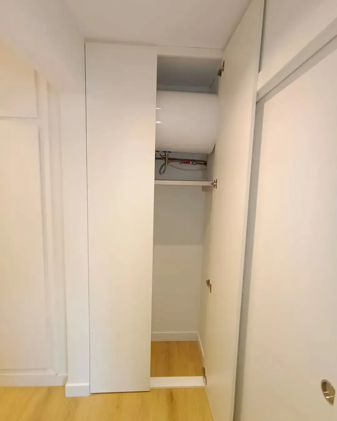 Armario blanco con puerta abierta, mostrando un estante y espacio de almacenamiento, piso de madera.