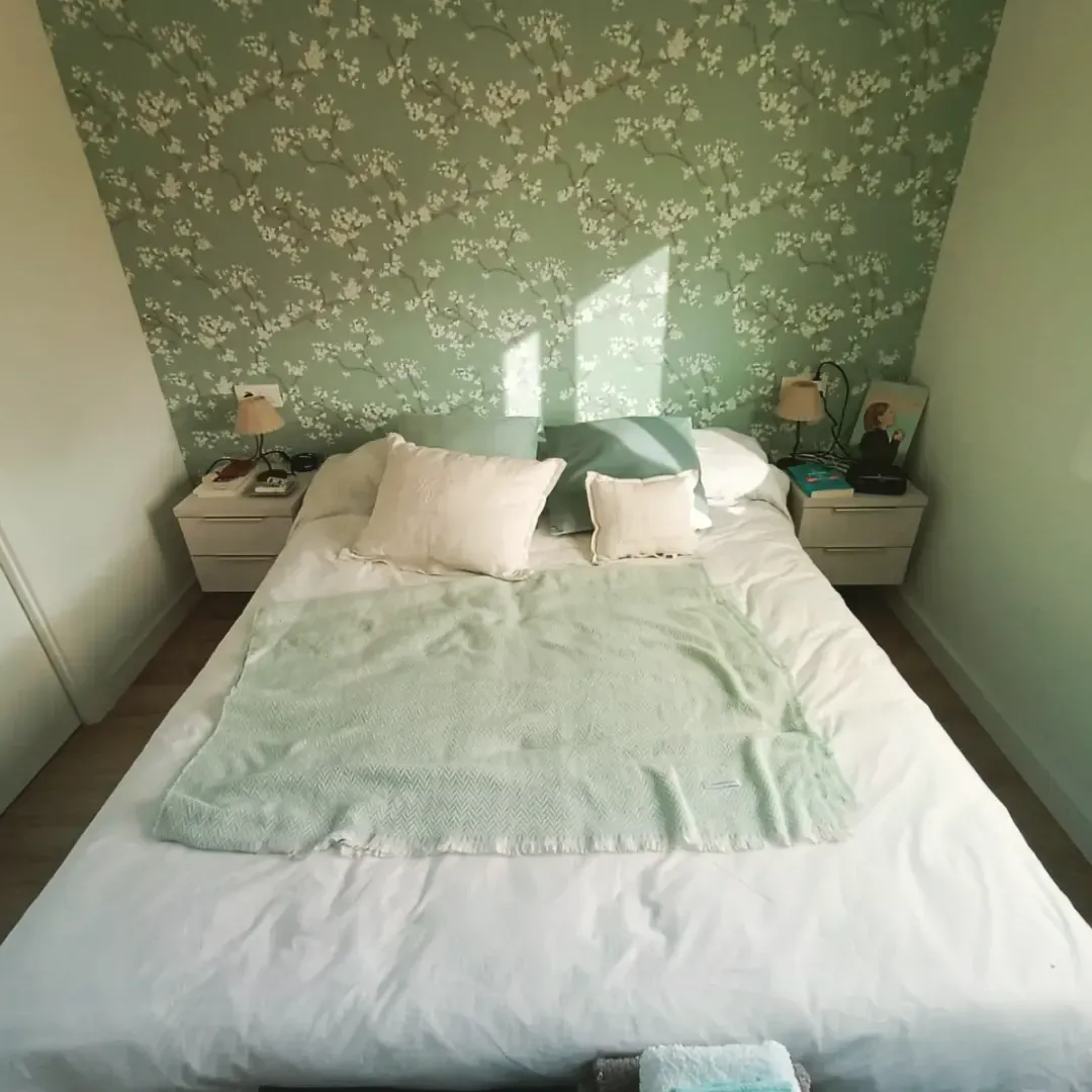 Dormitorio con cama blanca, manta verde y papel tapiz floral. Dos mesitas de noche con lámparas flanquean la cama.