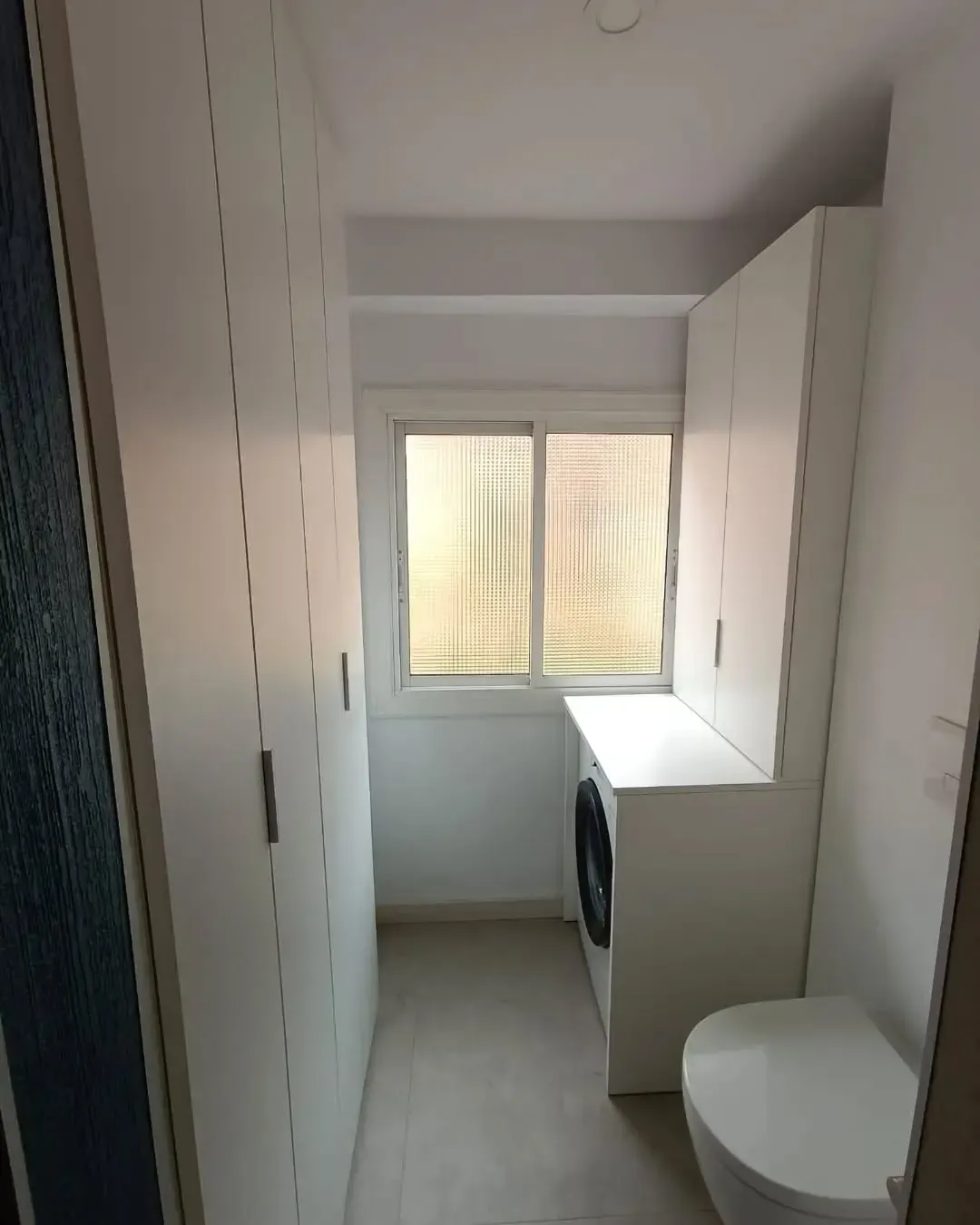Un lavadero estrecho con armarios blancos, lavadora, ventana y baño.