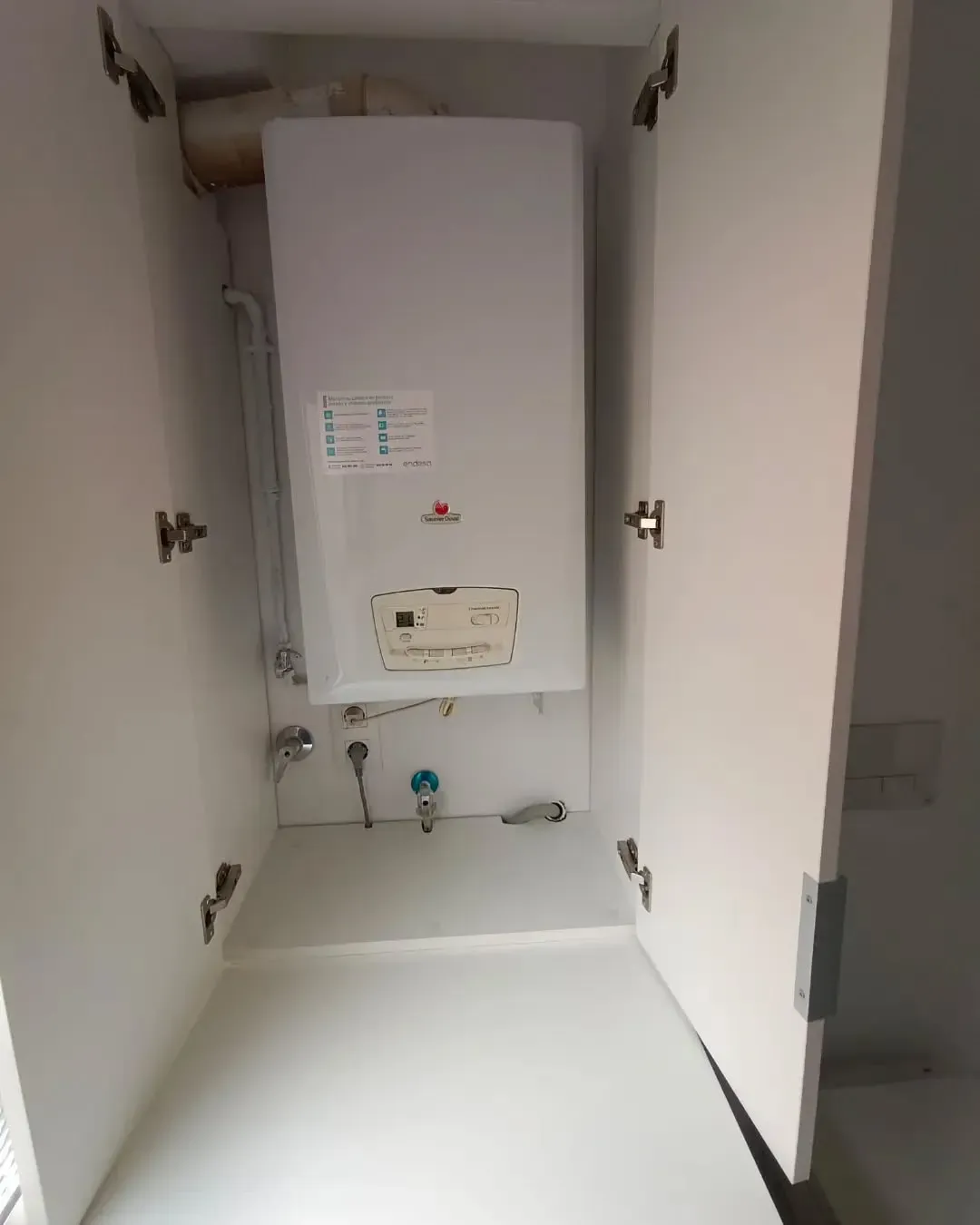 Caldera de gas blanca dentro de un armario blanco con las puertas abiertas. Tuberías, válvulas y etiquetas visibles.