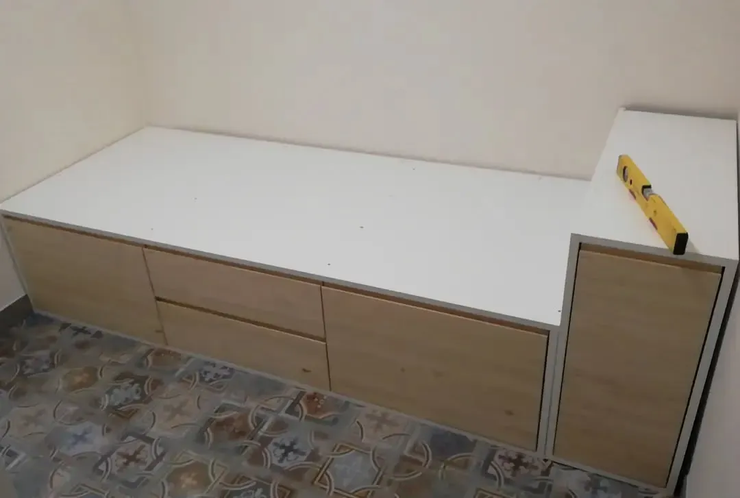 Cama empotrada con cajones de almacenaje y mesa auxiliar, madera clara y color blanco, un nivel reposa sobre la mesa.