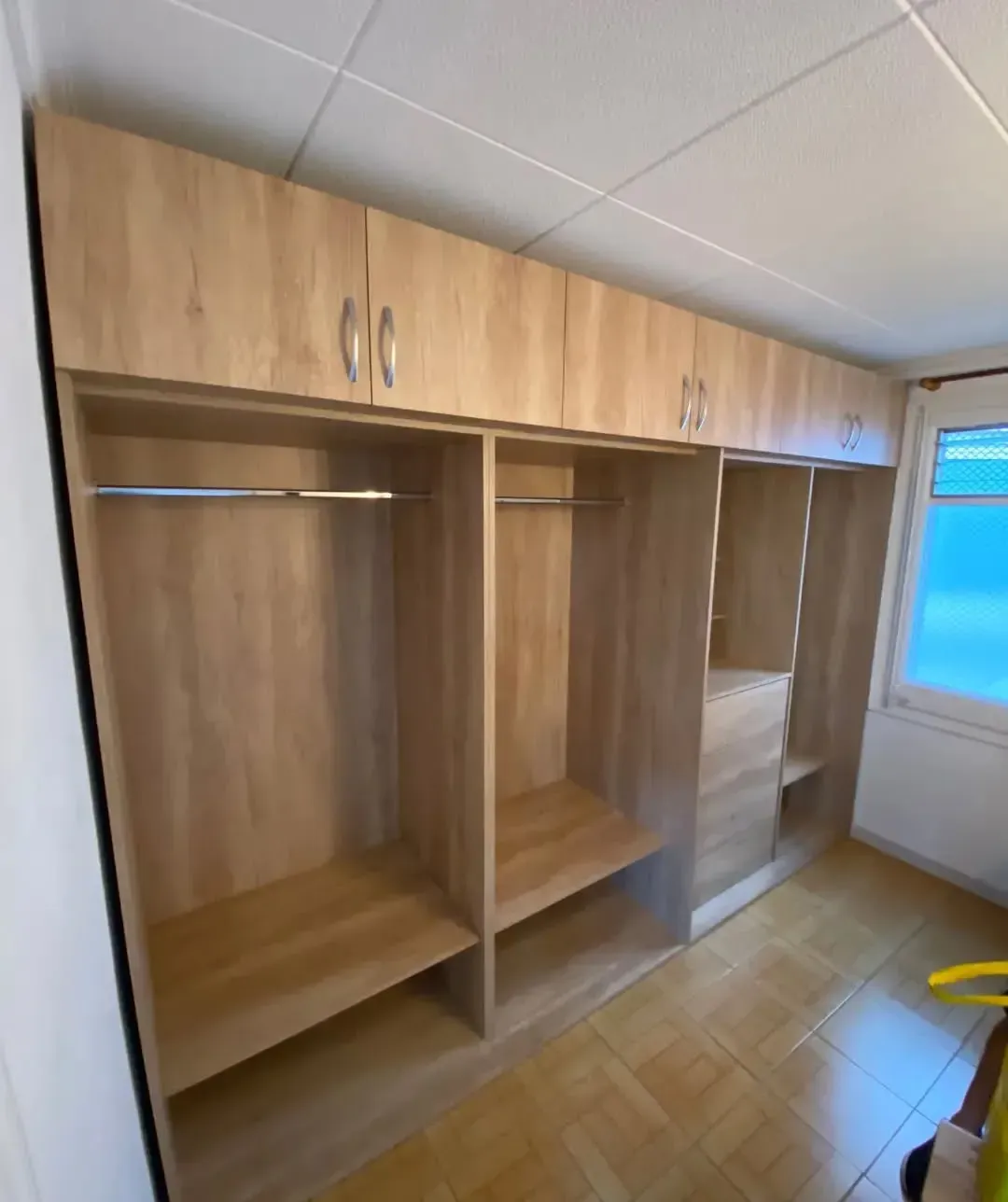 Armario empotrado de madera clara con barras para colgar, estantes y armarios. Suelo de baldosas claras.